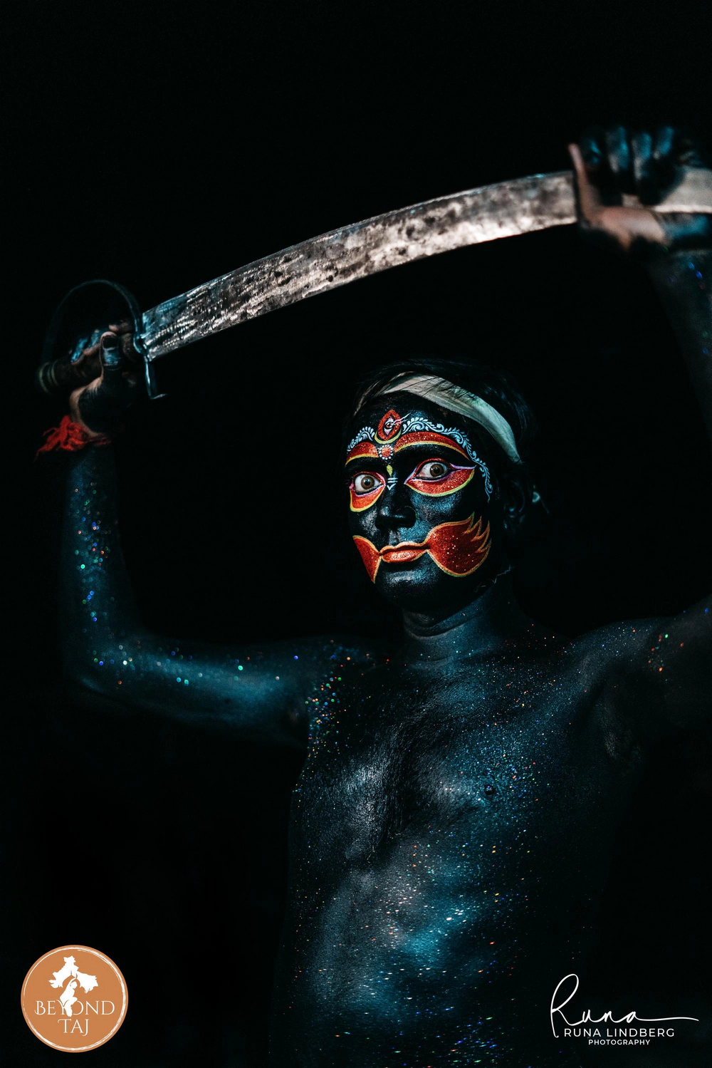 Painted entirely in black with red and gold markings across the face this Gajan performer raises a sword against total darkness. Poses like this are part of the dramatic deity performances staged throughout the festival nights.