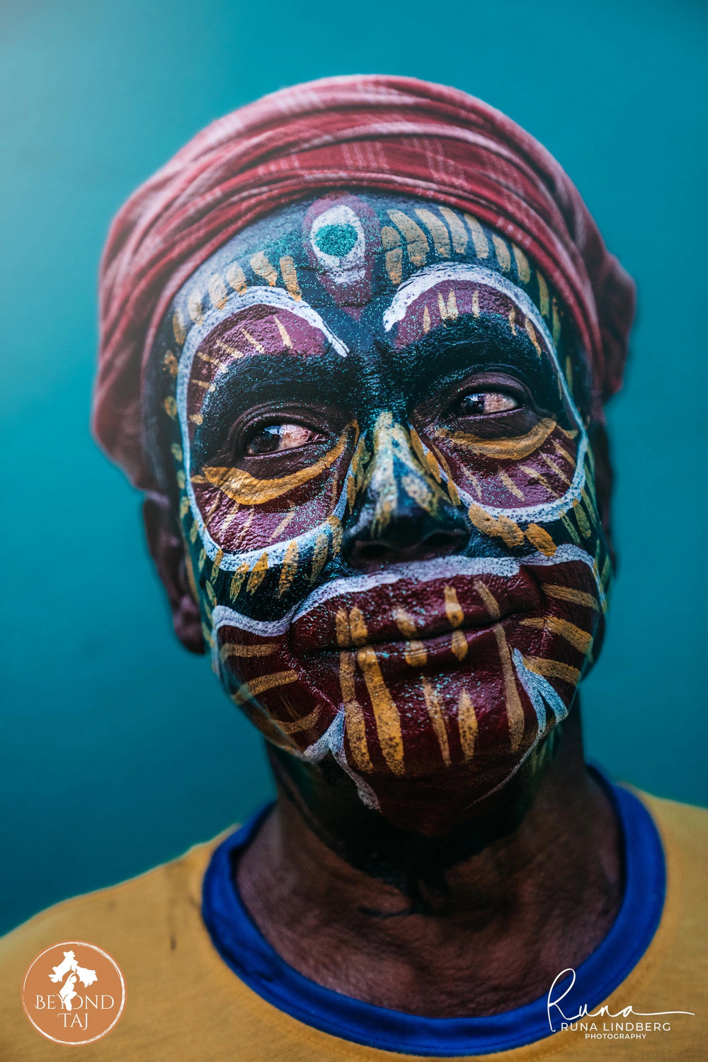 Fine stripes of maroon gold and white cover this devotees face in a tribal inspired pattern worn for Gajan. The red turban and cool teal backdrop set off the intricate handpainted design ahead of the nights procession.