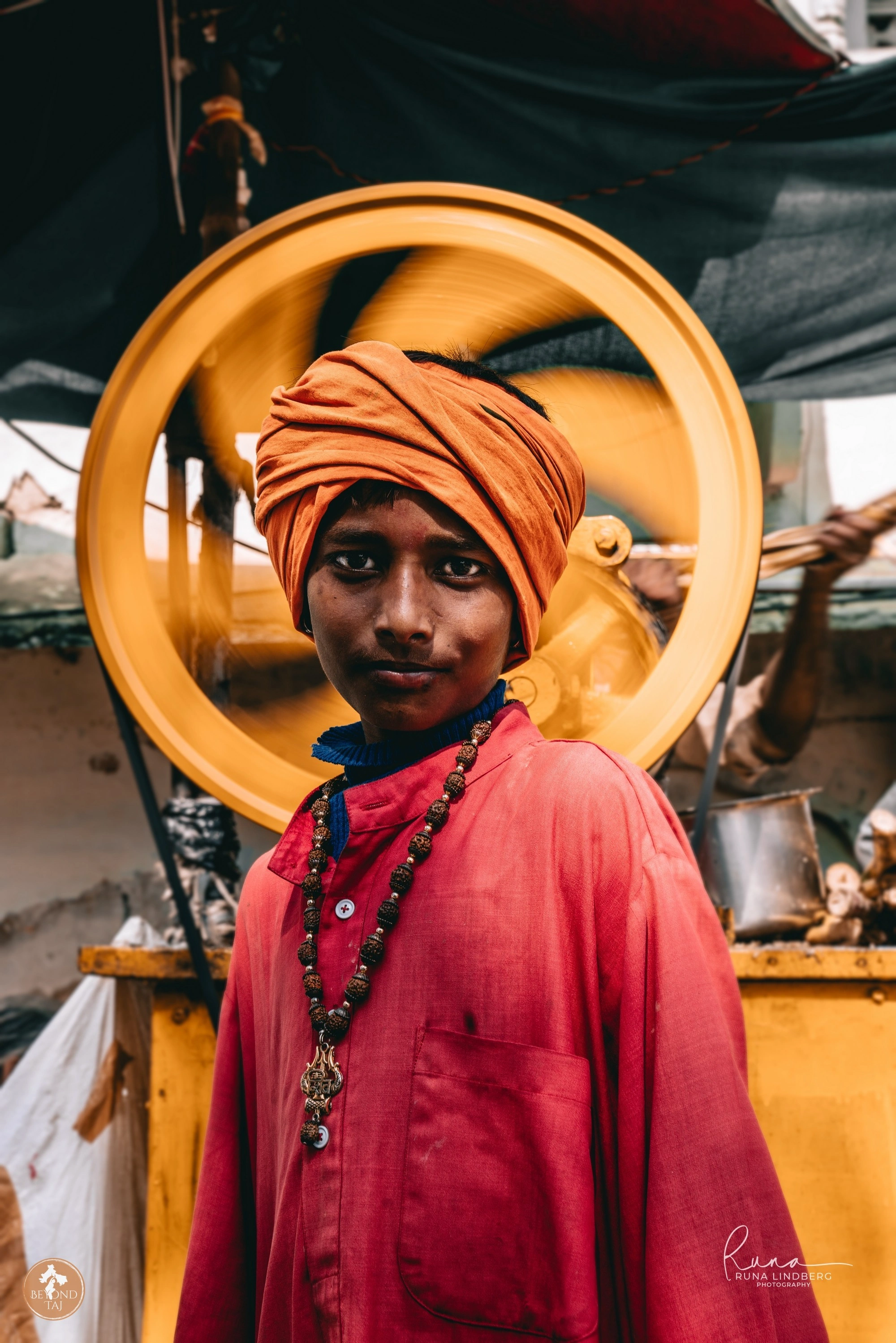 A young worker at the Pushkar Mela pauses in front of a spinning cart wheel his orange turban and rudraksha beads catching the afternoon sun. Fairground machinery like this keeps the camel carts and stalls running throughout the week.