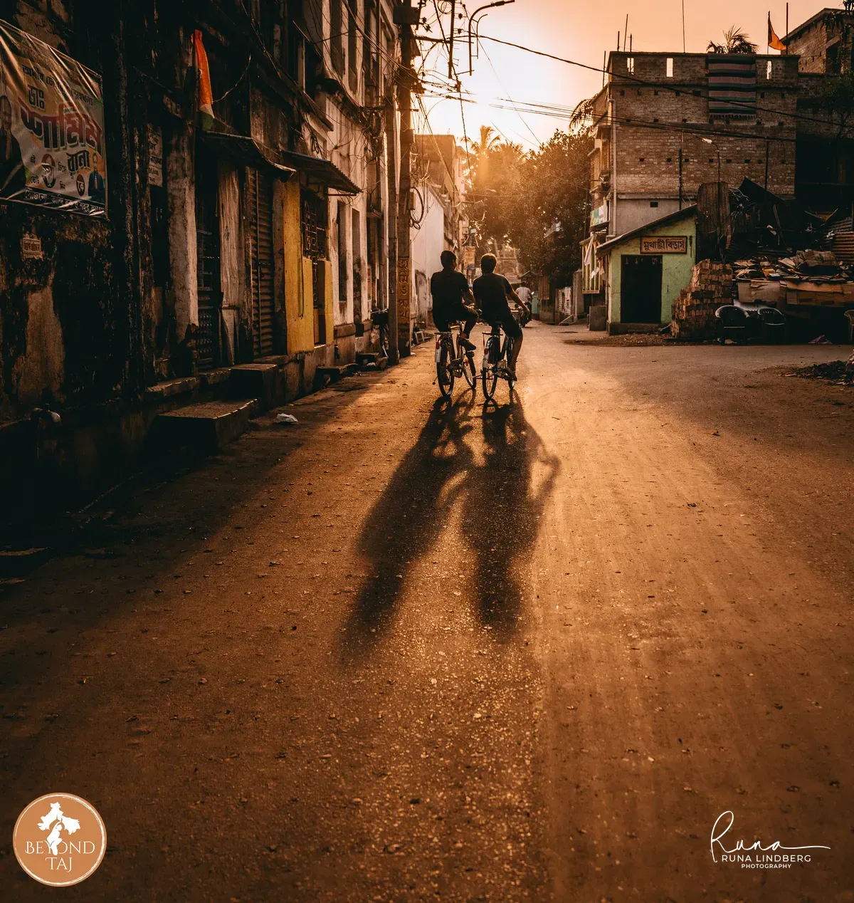 Two boys ride side by side through the streets of Kandi their long shadows trailing behind them in the low evening light. Faded shopfronts and hand painted signs line the road on either side.