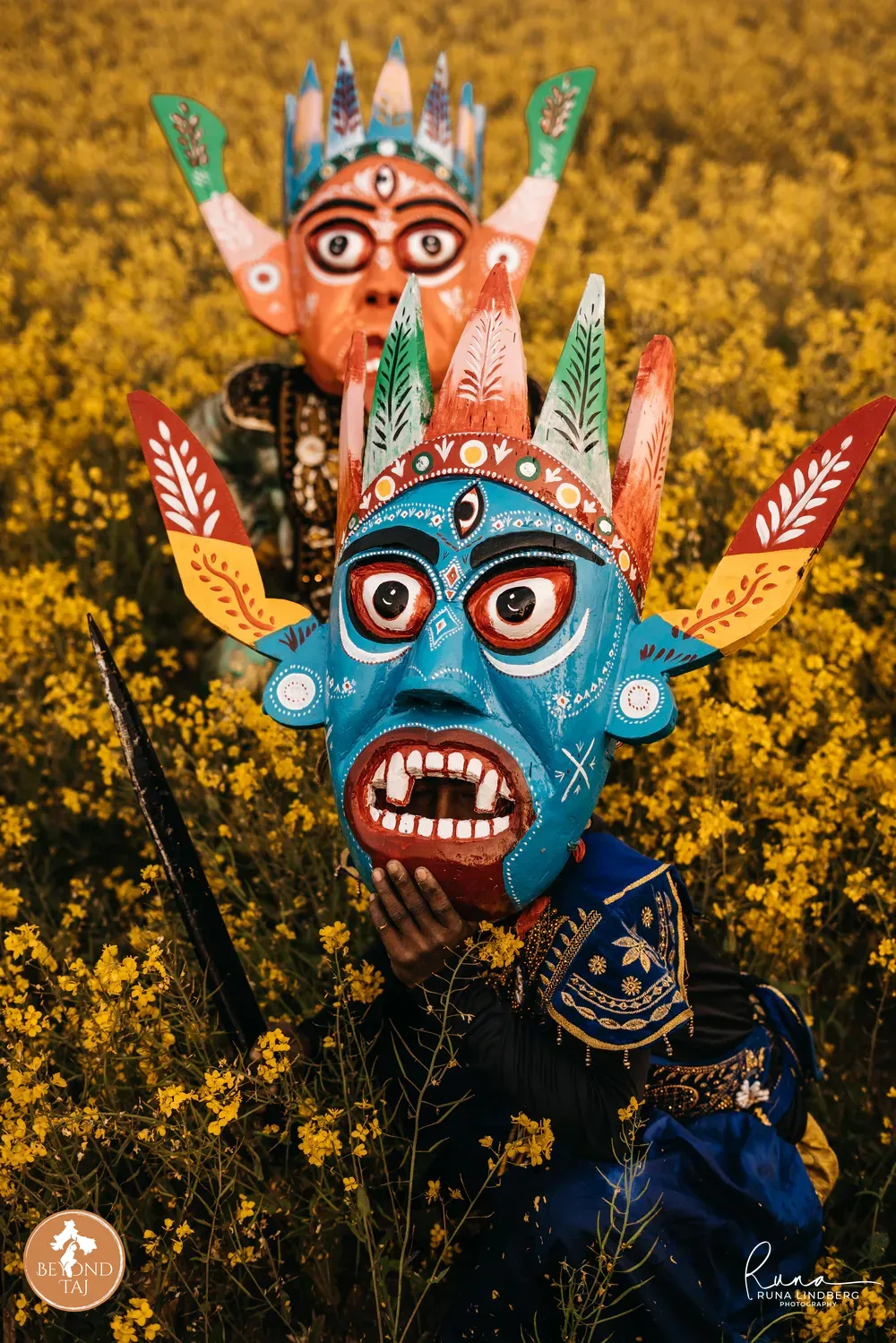 Two performers in ornate carved masks stand among the yellow blooms of a mustard field one holding a spear. Gomira dances are performed at dawn and dusk when the light and mist add to the dramatic effect of the costumes.