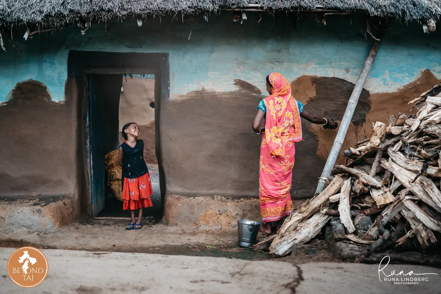 A woman smooths fresh mud plaster onto the wall of her home a maintenance task repeated regularly in rural Bengal. A young girl watches from the doorway smiling firewood stacked nearby for the coming season.