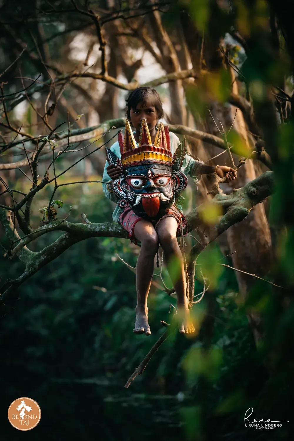 A girl perches on a tree branch cradling a red and gold Gomira mask in her lap her bare feet dangling below. The forest setting and quiet pose contrast with the fierce painted face of the mask she holds.