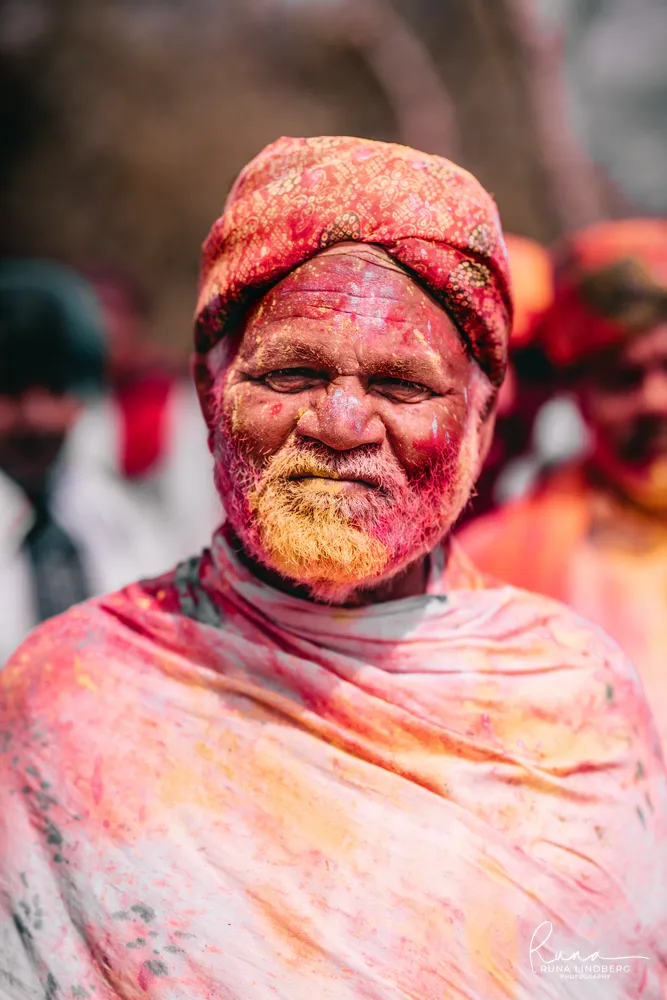 Portrait of an elderly man with a red patterned turban and white beard, his face and shawl covered in pink and yellow gulal, Holi in Braj