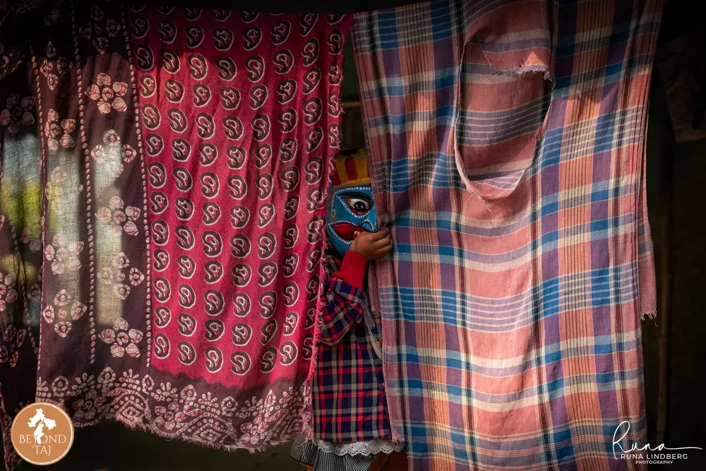 Young child peeking out from behind red scarves while wearing a Gomira mask, West Bengal