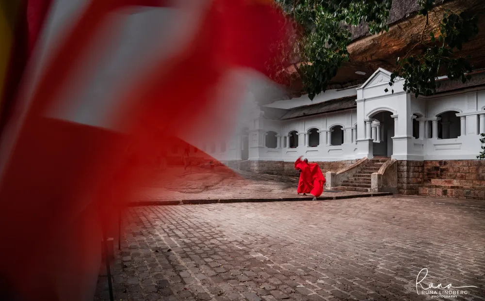 A monk in red robes crossing the temple courtyard at Dambulla, a prayer flag blurred in the foreground