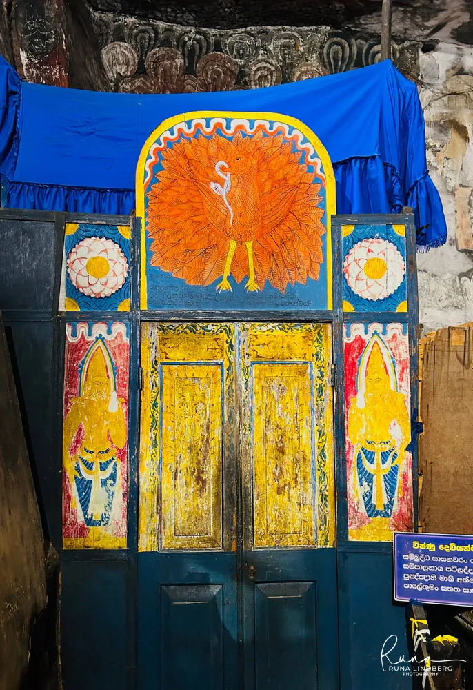 Painted wooden shrine doors at Dambulla topped by an orange peacock, with deity figures on the side panels