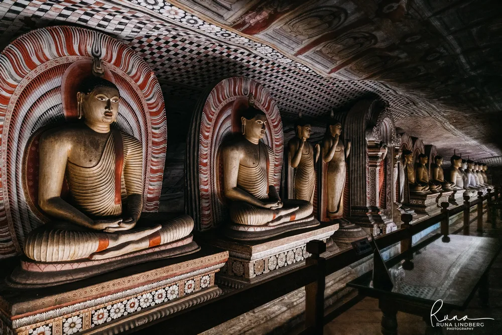 A long receding row of seated Buddha statues under a fully painted ceiling at Dambulla, each framed by a red and white arch