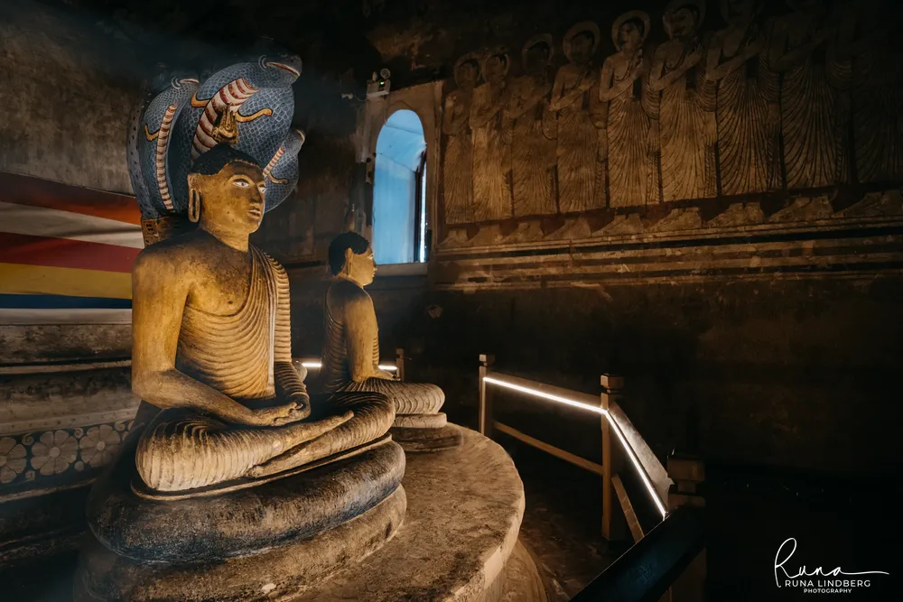 Seated Buddha sheltered by a multi-headed cobra hood in a dark cave shrine at Dambulla, lit by a small arched window