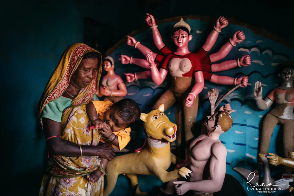 Woman in a yellow sari holding a baby in front of a partly painted clay Durga idol with ten arms, lion and demon, in a village in Purulia, West Bengal