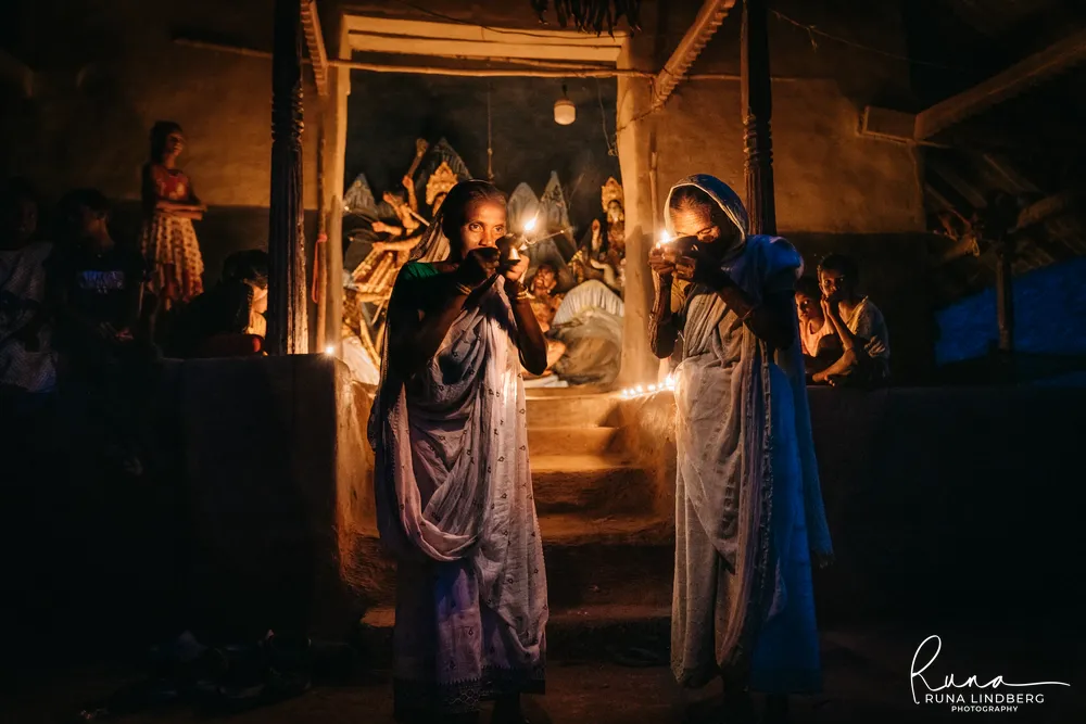 Two women in white saris holding small oil lamps in front of a village Durga shrine at night, children watching from the sides, Purulia, West Bengal