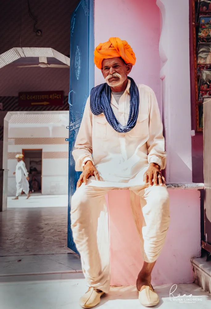 Elderly man in a bright orange turban, white clothes and a blue scarf sitting on a pink ledge beside a blue temple door, a sign behind him pointing to the Shri Devnarayan temple, Pushkar
