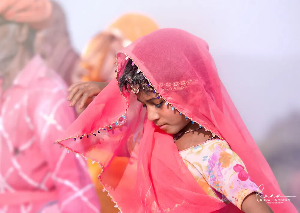 Young girl in a sheer pink veil edged with sequins looking down, family members in bright clothes behind her, Pushkar