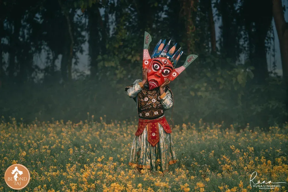 Gomira dancer in a carved wooden mask crossing a mustard field in full yellow bloom, Kushmandi, Dakshin Dinajpur, West Bengal