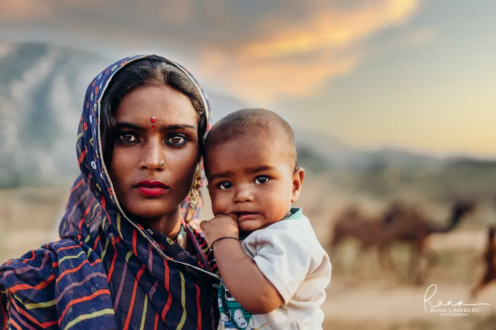 Young woman in a striped blue veil holding a small child, camels blurred in the background at sunset on the dunes, Pushkar