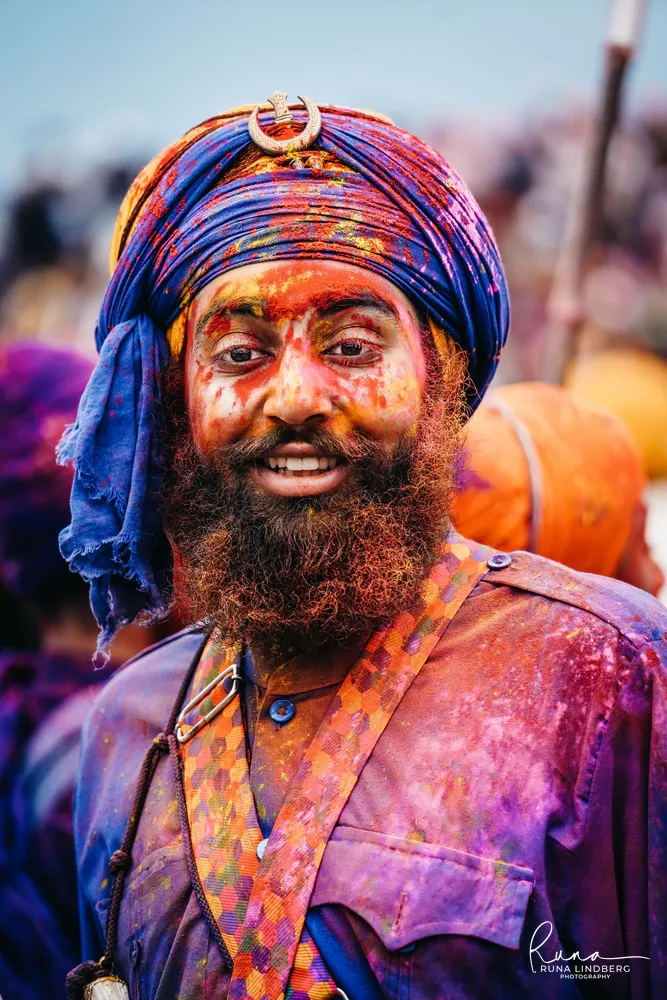 Smiling Nihang Sikh in a blue turban, his face and beard covered in red, yellow and purple colour powder, Hola Mohalla, Anandpur Sahib