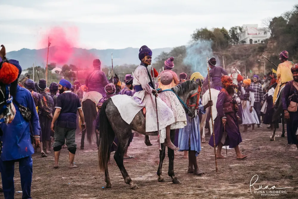 Nihang riders on horseback gathering on open ground while clouds of pink and blue colour powder rise behind them, Hola Mohalla, Anandpur Sahib
