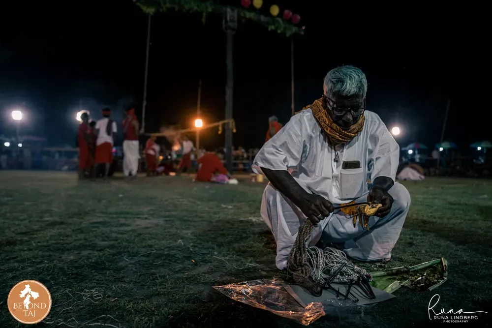 a man in white kneeling on grass at night, handling rope and metal hooks, string lights behind