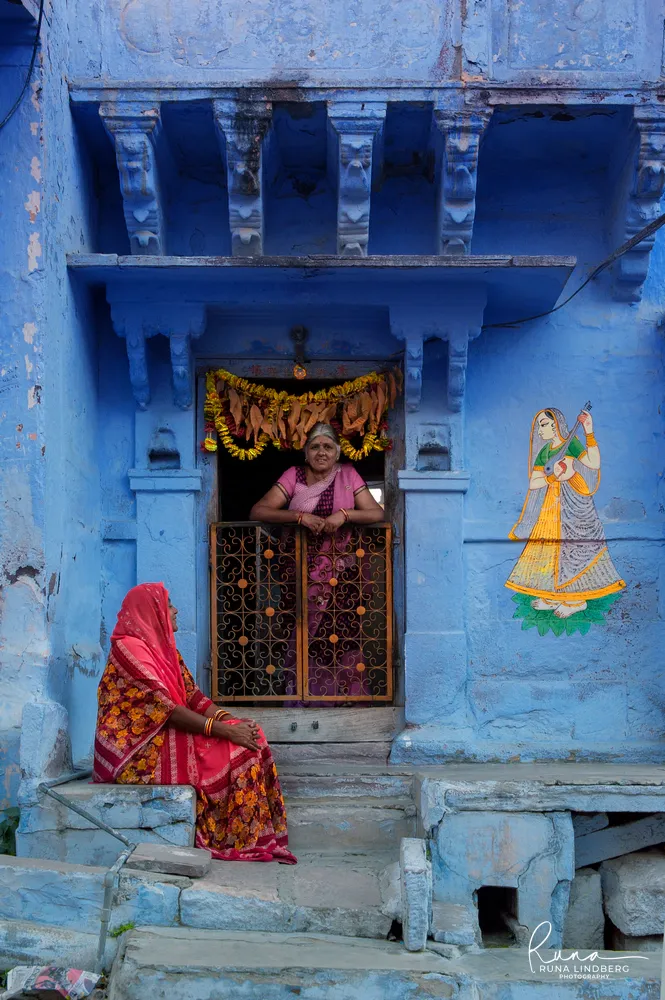 Two women at a blue house in Jodhpur's old city, one leaning on a wrought-iron half-door under a marigold garland, one in a red sari sitting on the steps, a painted female figure on the wall