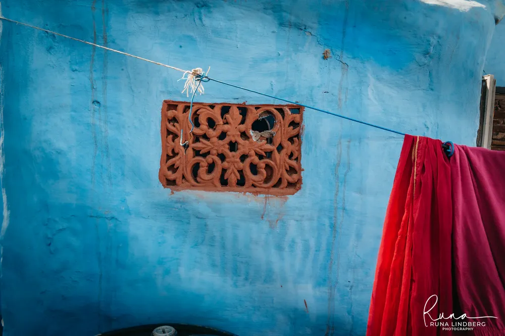 Terracotta-coloured carved window grille in a blue wall in Jodhpur's old city, a washing line with red cloth running across it