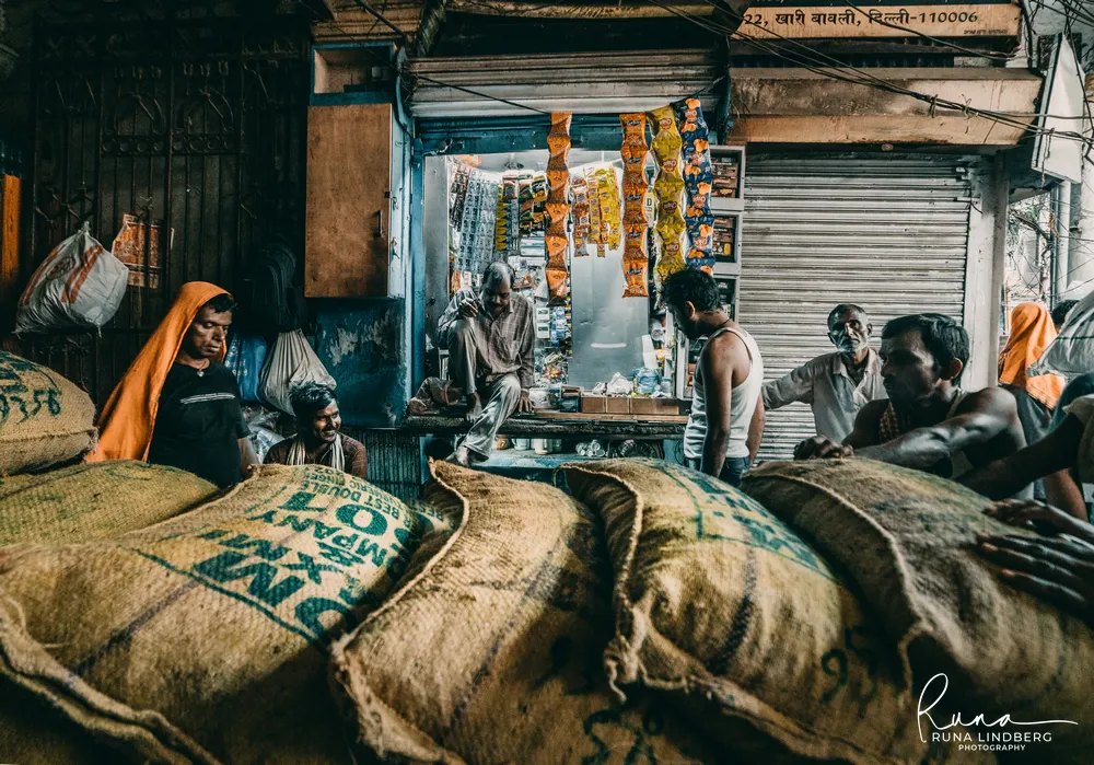Porters resting among stacked jute sacks in front of a small kiosk hung with snack packets in Khari Baoli, Old Delhi