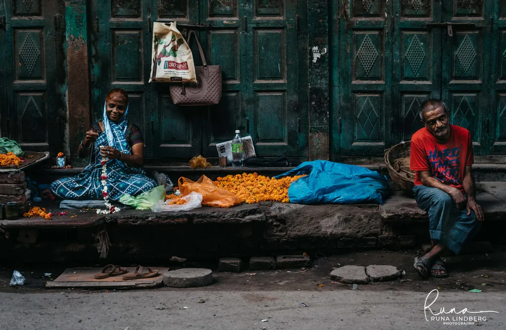 Woman in a blue checked sari threading a flower garland on a stone ledge in front of old green wooden doors, marigolds piled beside her, a man in a red T-shirt sitting to the right, Old Delhi
