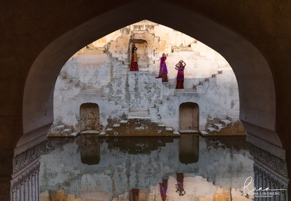 Three women in red and purple saris with water pots on their heads on the zigzag steps of Panna Meena ka Kund stepwell in Amer, framed by a dark stone arch and mirrored in the water