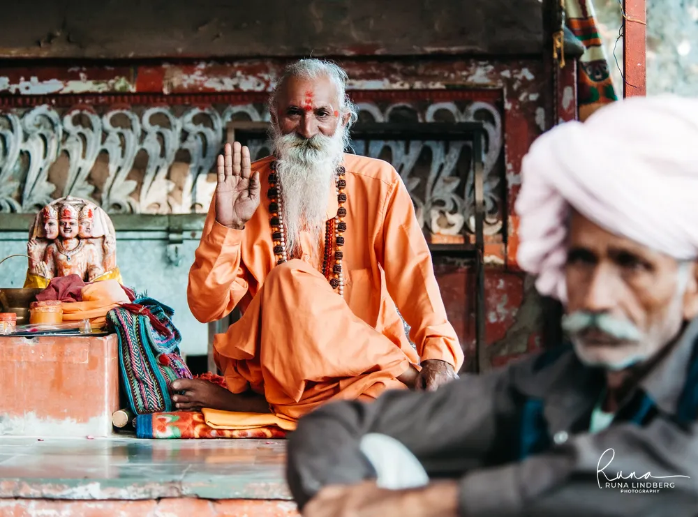 Sadhu in saffron robes with rudraksha beads raising his hand in blessing beside a small shrine with a figure of the four-faced god Brahma, an elderly man in a pink turban in the foreground, Pushkar