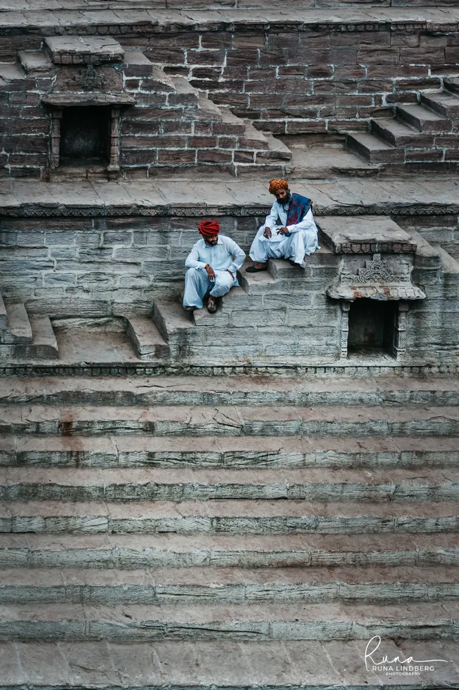 Two men in white with red and orange turbans sitting on the stone steps of Toorji ka Jhalra stepwell in Jodhpur, seen from above