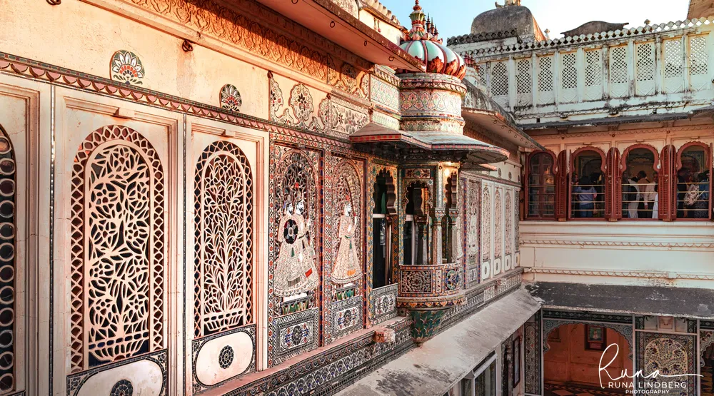 Upper storey of a courtyard at Udaipur City Palace with carved jali screens, glass-mosaic figures and a projecting balcony under a red and white dome