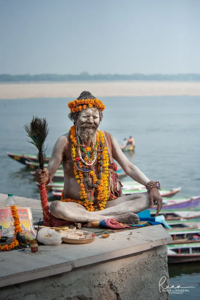 Ash-covered sadhu with marigold garlands and prayer beads sitting cross-legged on a platform above the Ganges in Varanasi, a peacock-feather fan in his hand, boats moored below