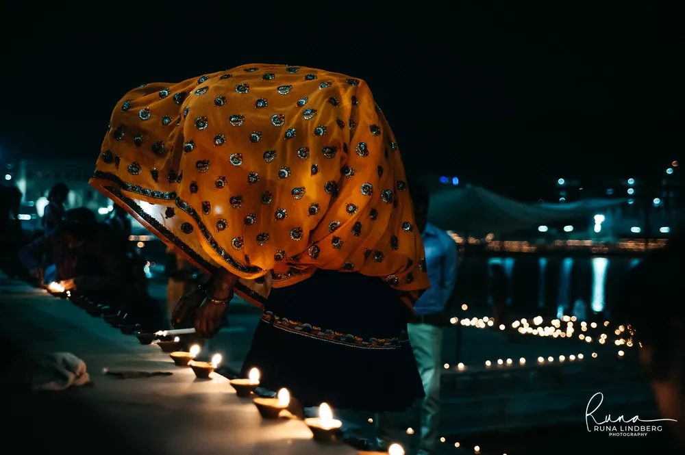 Woman in a yellow embroidered veil bending to light oil lamps along the edge of a ghat at night, lights reflected in Pushkar Lake