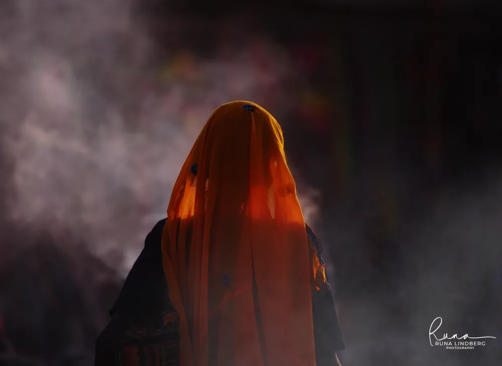 Woman with an orange veil drawn over her face on her way to the herders' camp, backlit by the sun through drifting smoke, Pushkar Mela
